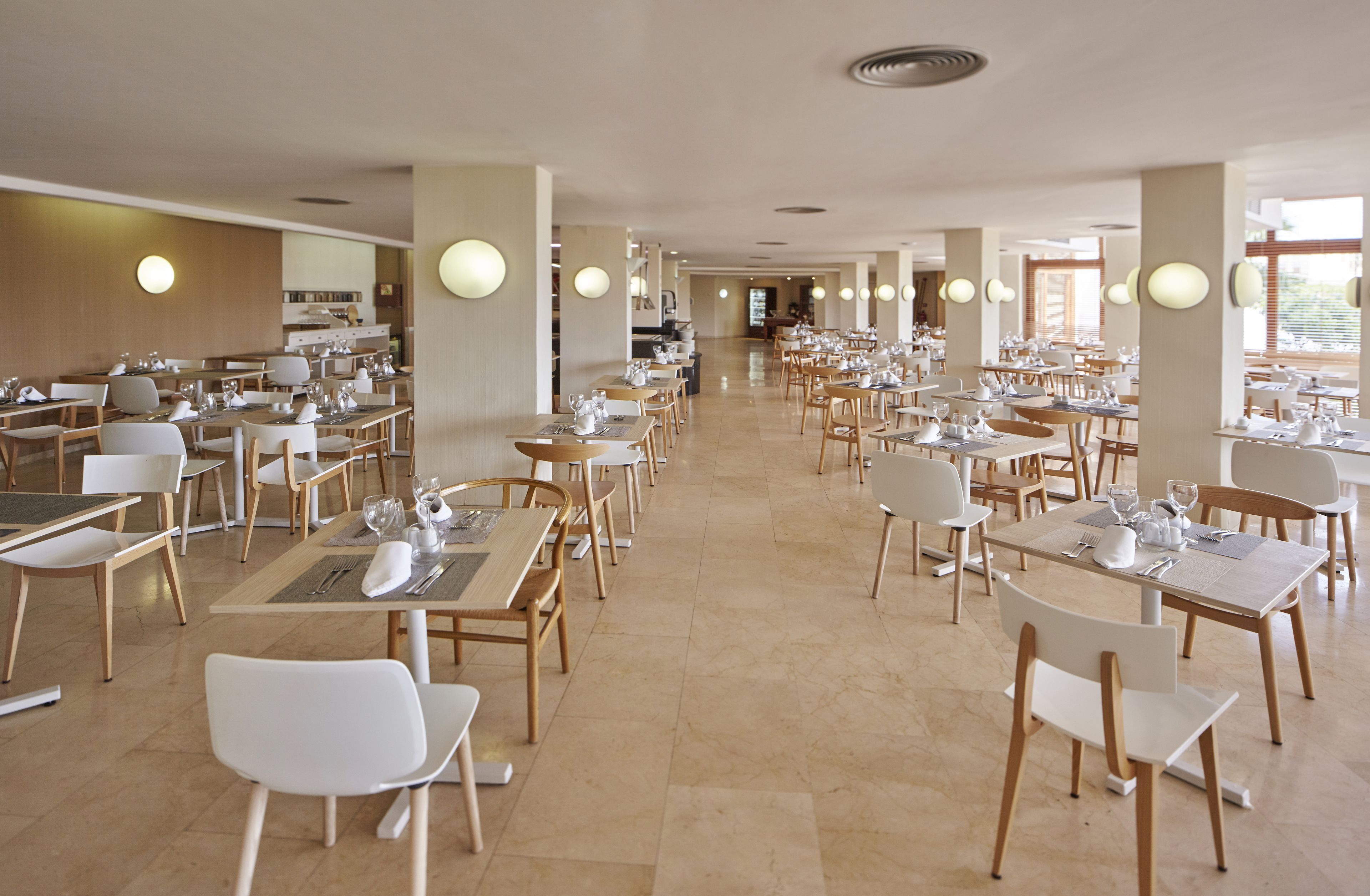 A spacious dining area with multiple tables and chairs, arranged in rows, and a kitchen area in the background.