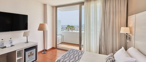 A hotel room with a bed, a TV, a desk with a lamp, and a view of the sea.
