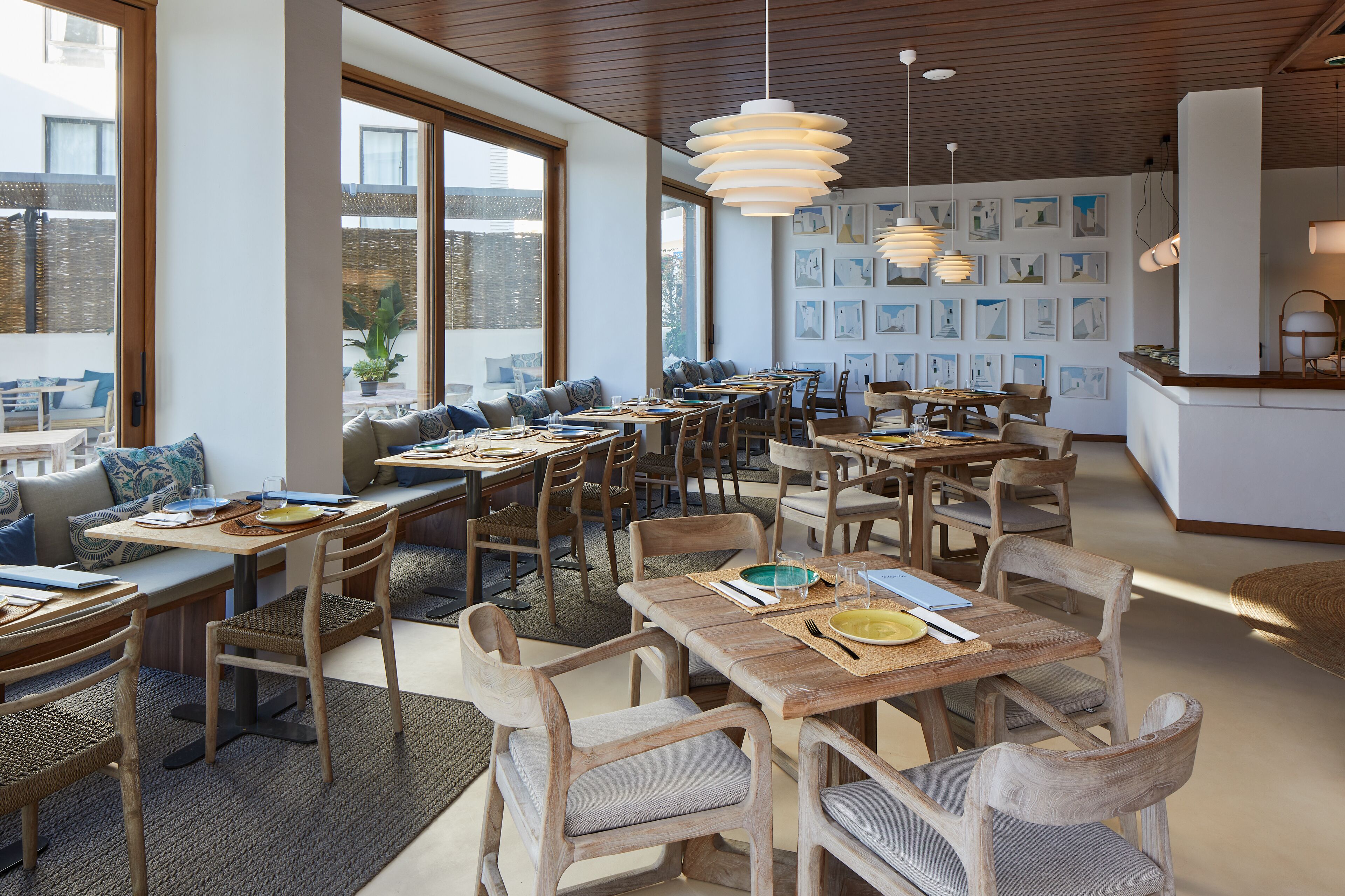 A modern dining area with wooden tables and chairs, large windows, and artwork on the walls.