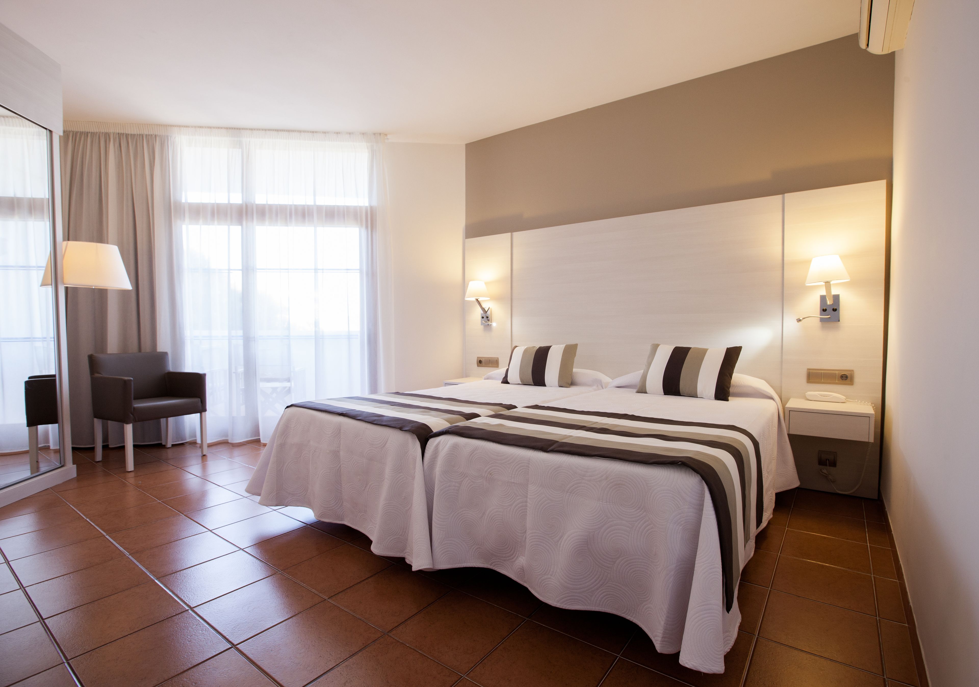 A hotel room with a large bed, two bedside lamps, a chair, and a window with curtains.