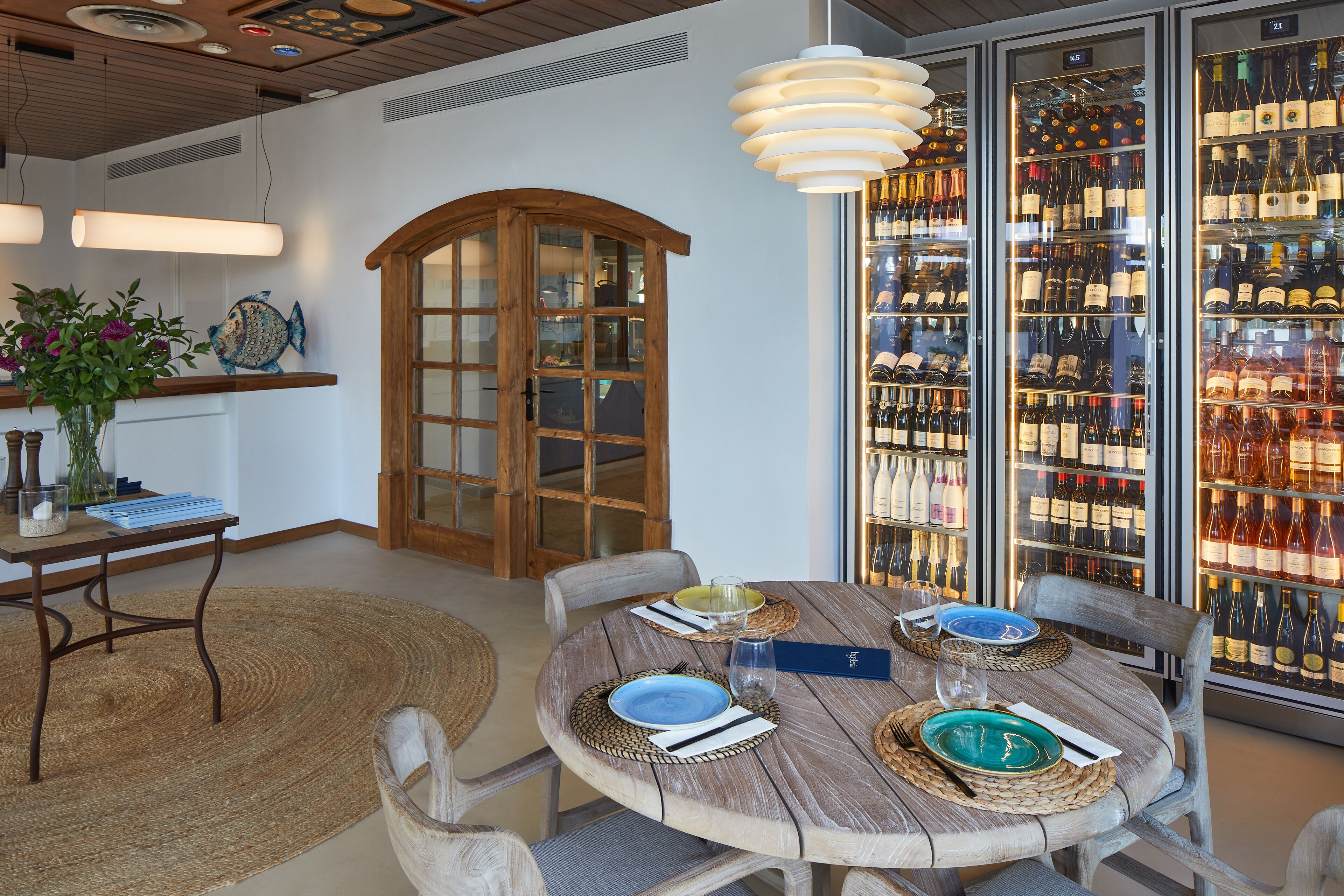 Uma área de refeições com uma mesa redonda de madeira posta para quatro pessoas, rodeada por cadeiras. Uma adega grande é visível ao fundo, com diversas garrafas de vinho.
