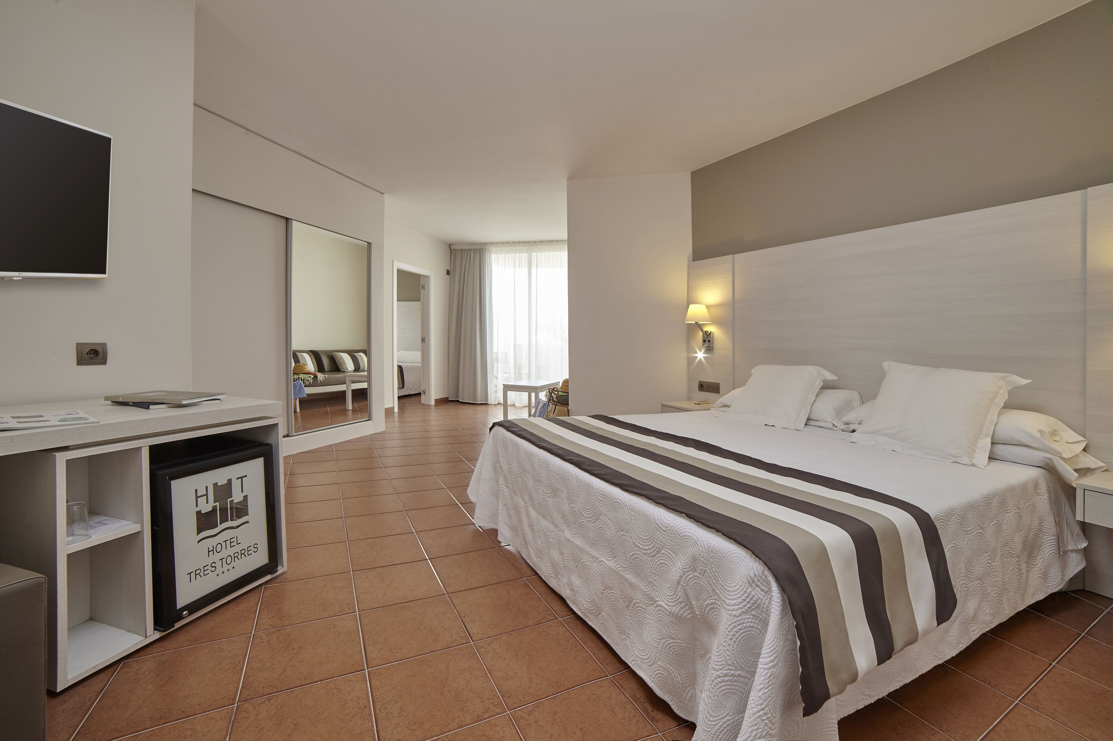 A hotel room with a bed, bedside table, lamp, TV, and a desk with a computer and papers.