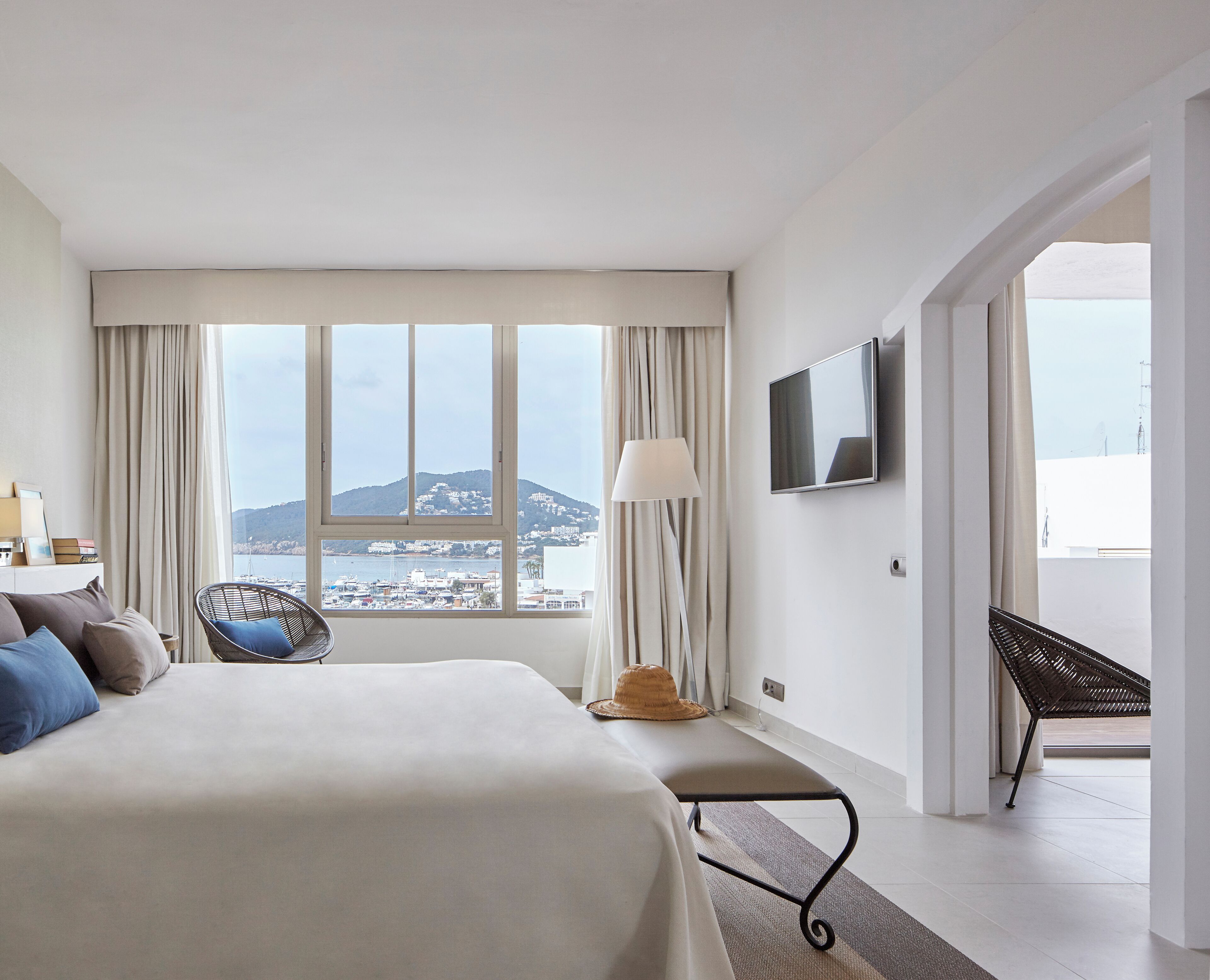 A hotel room with a large bed, a chair, a TV, and a view of a coastal town.