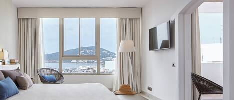 A hotel room with a large bed, a chair, a TV, and a view of a coastal town.