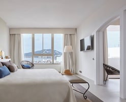 Et hotelværelse med en stor seng, en stol, et fjernsyn og udsigt over en kystby.