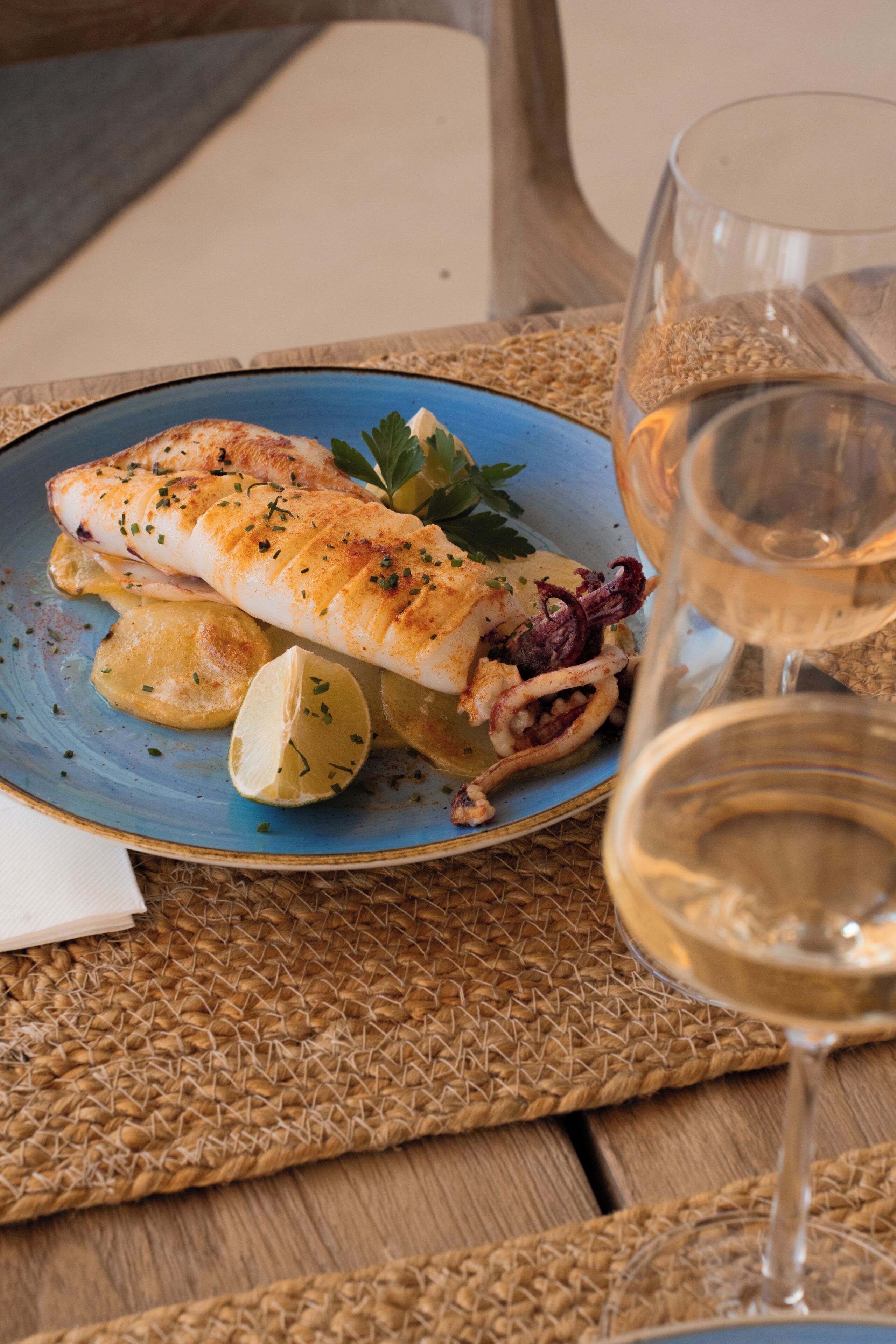 Un plato de pescado con limón y hierbas, una copa de vino blanco y un posavasos tejido.