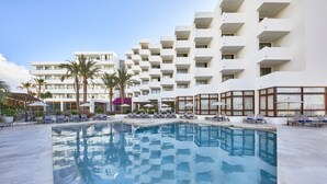 Zona de piscina con sillas de descanso y palmeras, rodeada por un edificio de varios pisos con balcones.
