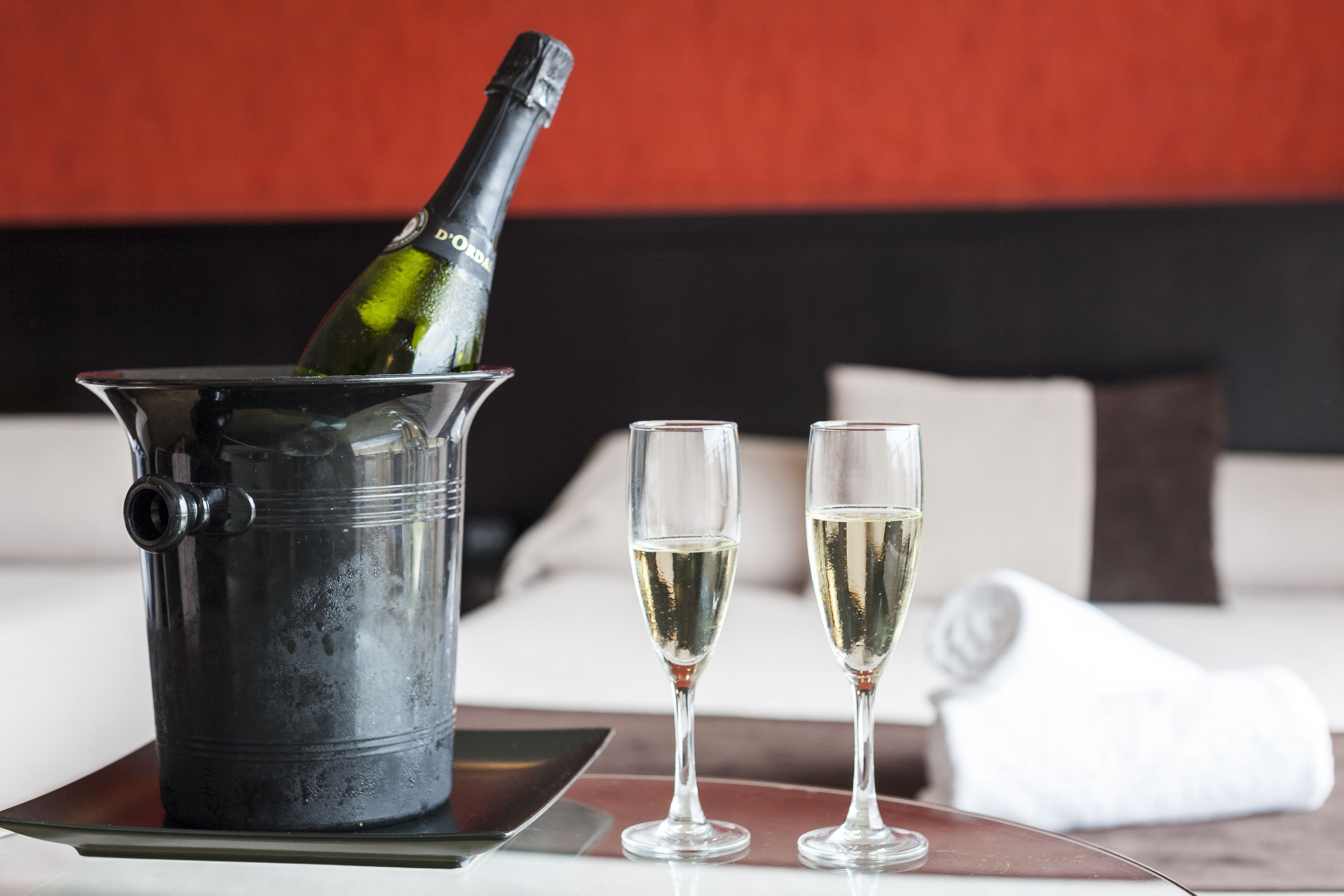 A bottle of champagne in a metal bucket, two glasses of champagne, and a white towel on a table in a room with a red and black wall.