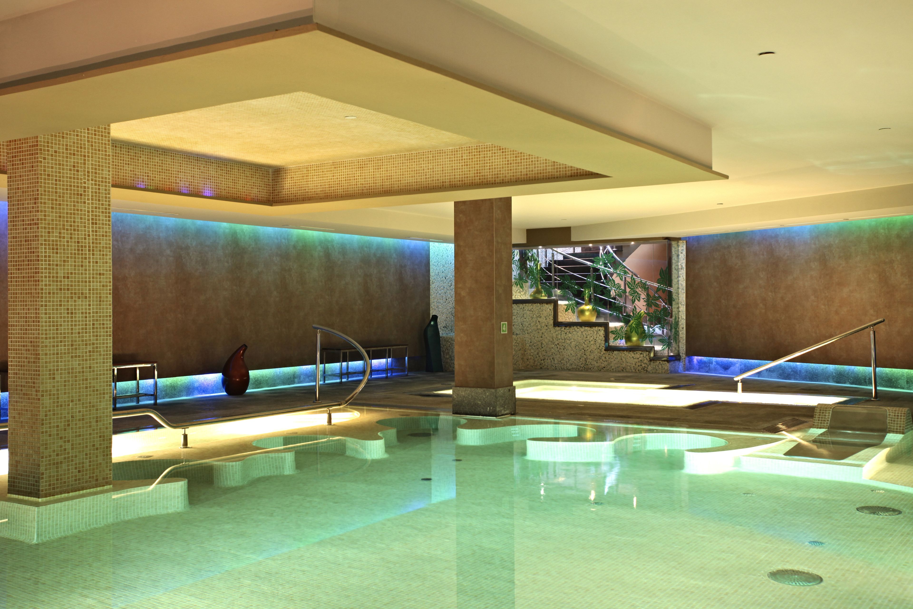 A large indoor swimming pool with multiple lanes, a central water feature, and a seating area with a staircase leading to an upper level.