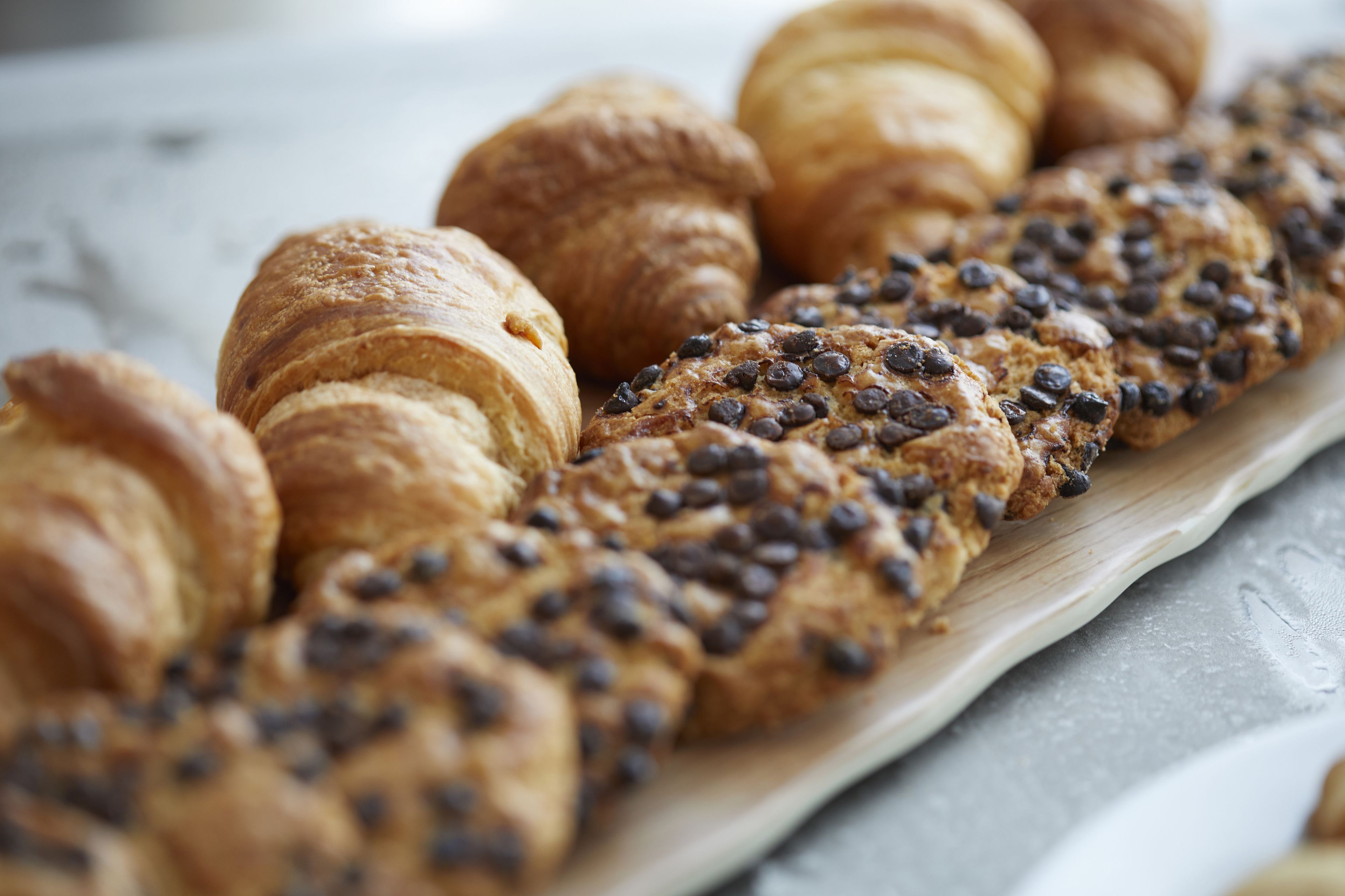 A variety of baked goods, including croissants and a chocolate chip cookie bread, displayed on a wooden board.