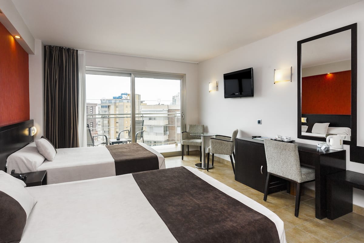 A hotel room with two beds, a desk, a chair, a TV, and a large window with a city view.