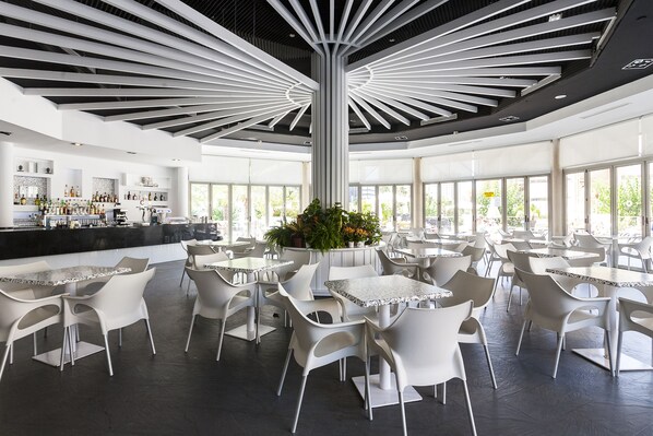 A spacious indoor area with white tables and chairs, a central column, and a bar area with shelves of bottles.