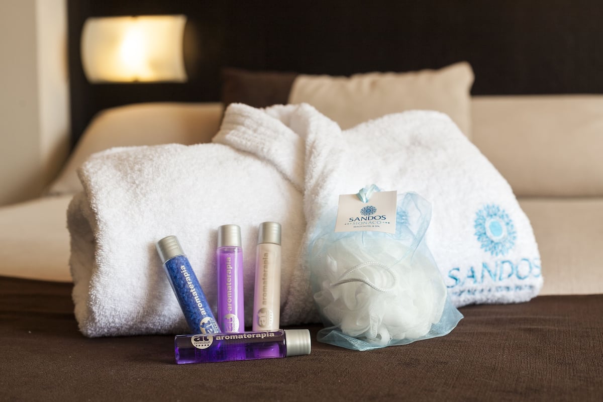 A hotel room with a bed, a white towel, a bag of white bath products, and three bottles of bath oils.