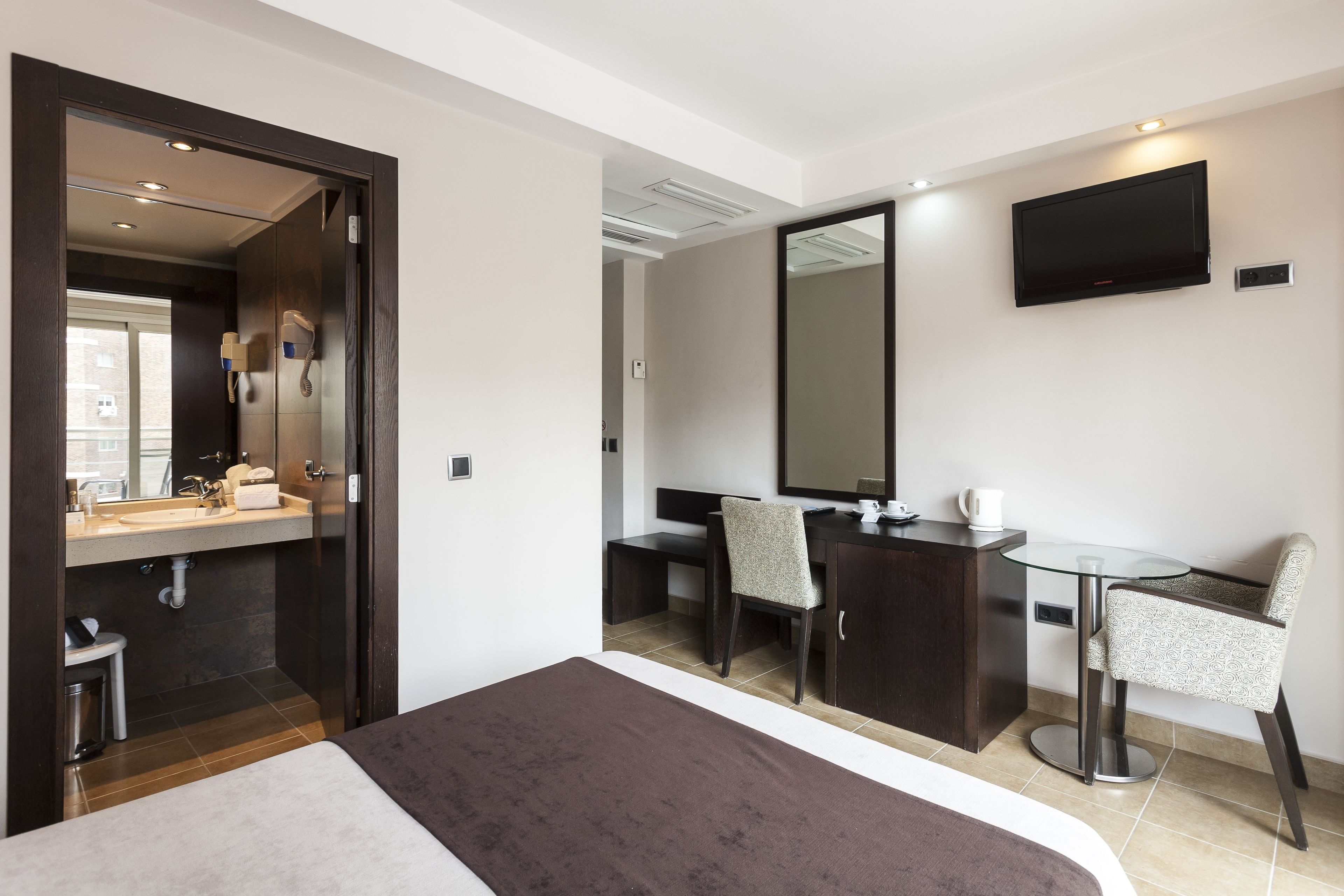 A modern hotel room with a bed, a desk, a chair, a large mirror, and a TV on the wall.