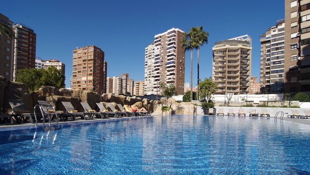Una piscina con sillones de descanso y palmeras, rodeada de edificios altos.