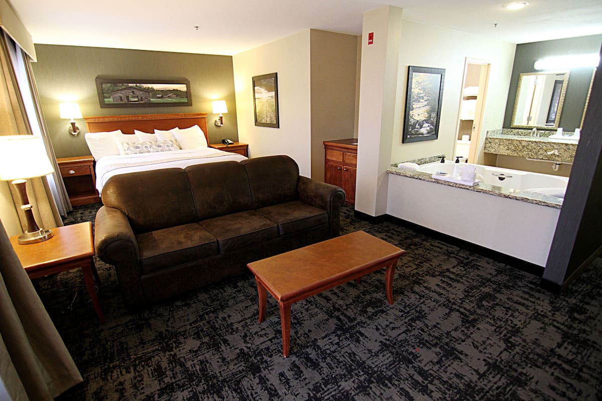 A hotel room with a bed, a sofa, a small table, a bathroom with a sink and mirror, and a nightstand with a lamp.
