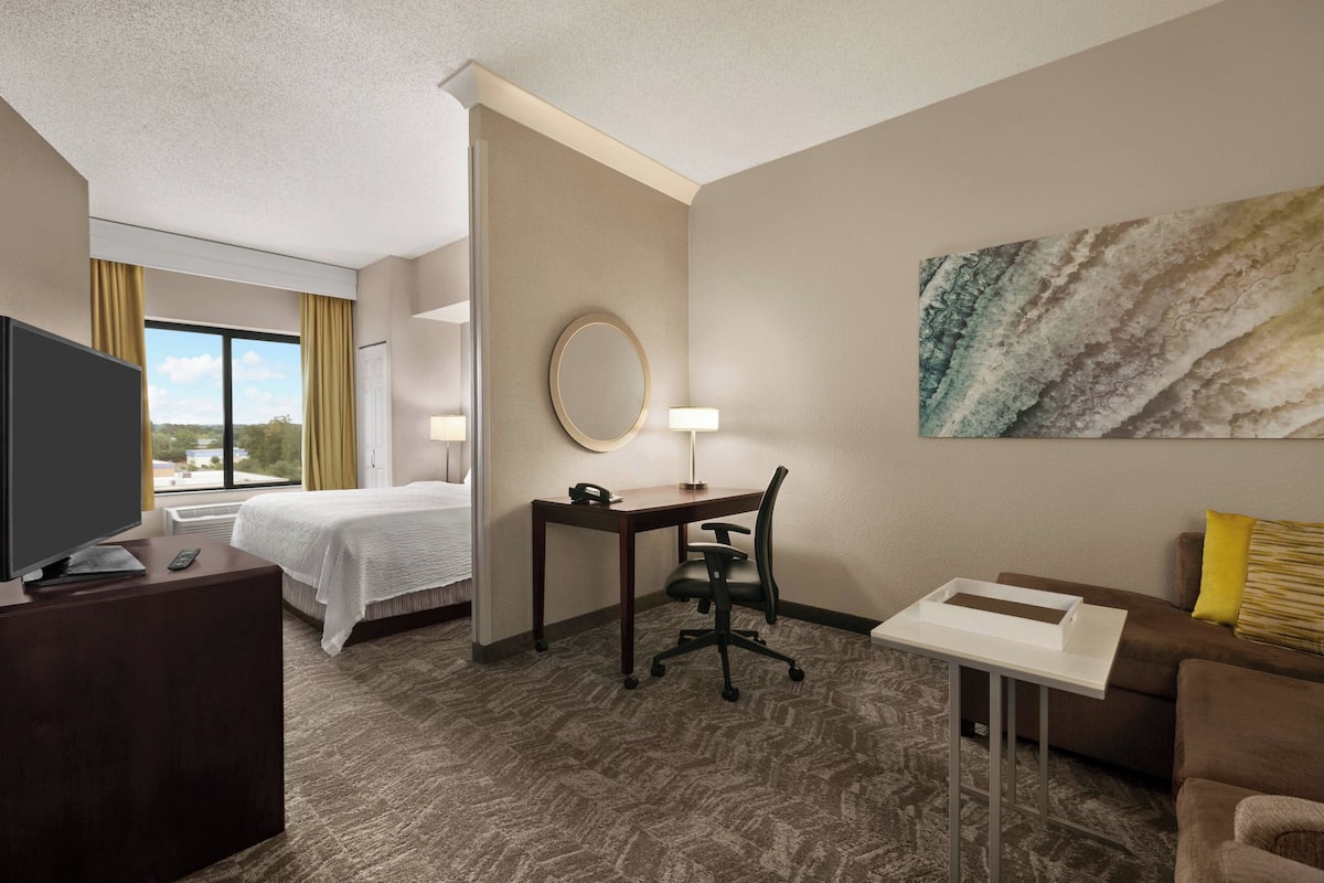 A hotel room with a bed, desk, chair, TV, and a painting on the wall.