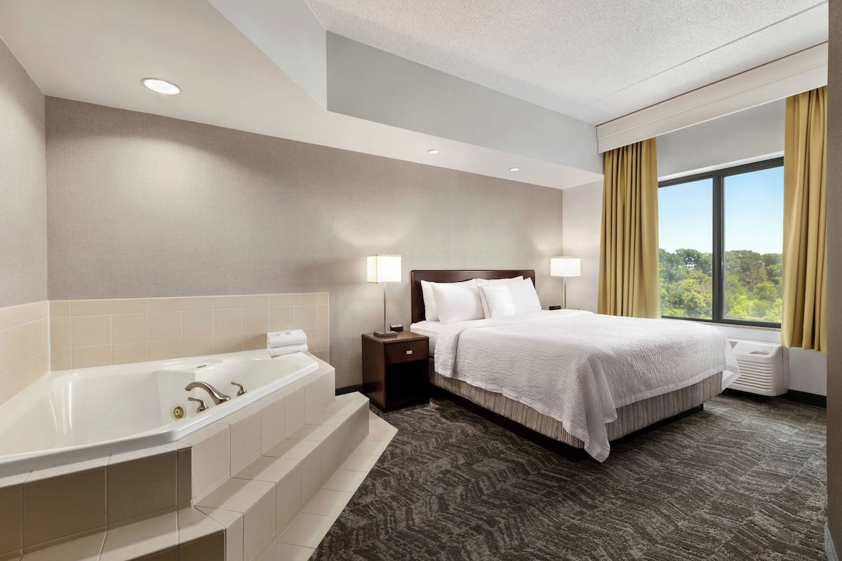 A hotel room with a large bathtub, a bed with white linens, two bedside lamps, and a window with curtains.