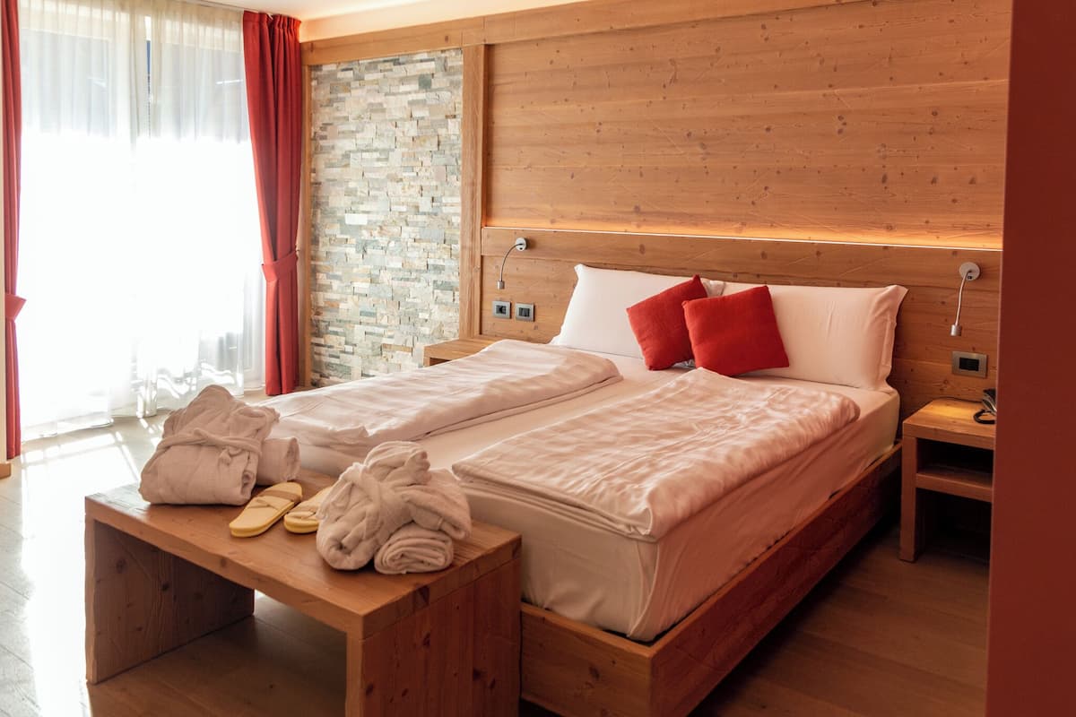 Een tweepersoonsbed met houten hoofdbord, een bankje met handdoeken en slippers, en een stenen muur.
