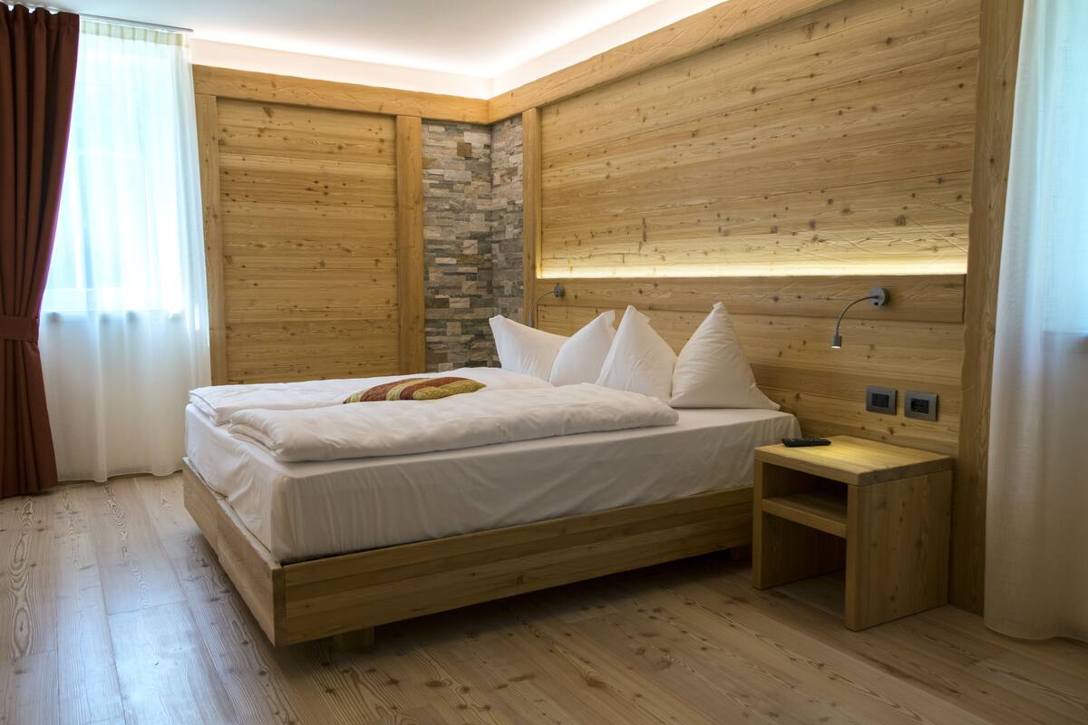 Een bed met houten frame, witte beddengoed en kussens, een nachtkastje met een lamp en een accentmuur van steen.