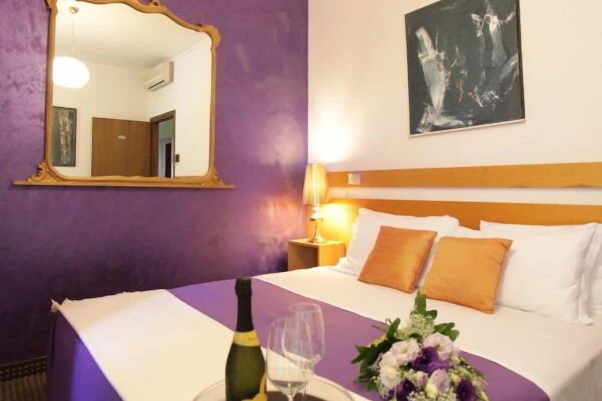 A hotel room with a purple wall, a large mirror, a bed with white bedding, a nightstand with a lamp, a bottle of champagne, a glass, and a floral arrangement.