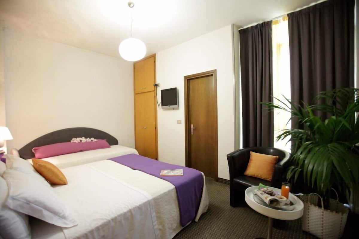 A hotel room with a bed, a chair, a small table with a tray, a television, and a large plant.