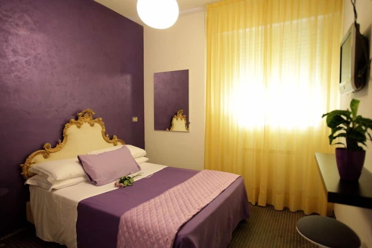A hotel room with a bed, a mirror, a window with yellow curtains, and a potted plant.