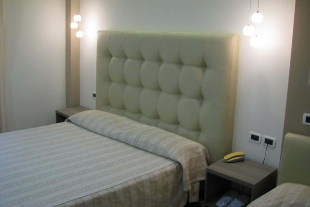 Un dormitorio con cabecera capitonada, cama con colcha a rayas, mesita de noche con lámpara y velador con teléfono.