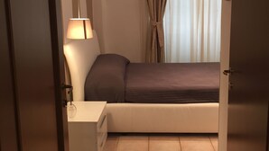 Quarto compacto com cama, mesa de cabeceira com luminária, escrivaninha pequena e janela com cortinas.