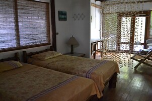 Una habitación con dos camas, piso de madera, ventana con persianas, una lámpara y una silla.