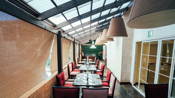 Un restaurante con zona de estar exterior cubierta, sillas tapizadas rojas y una pared verde al fondo.