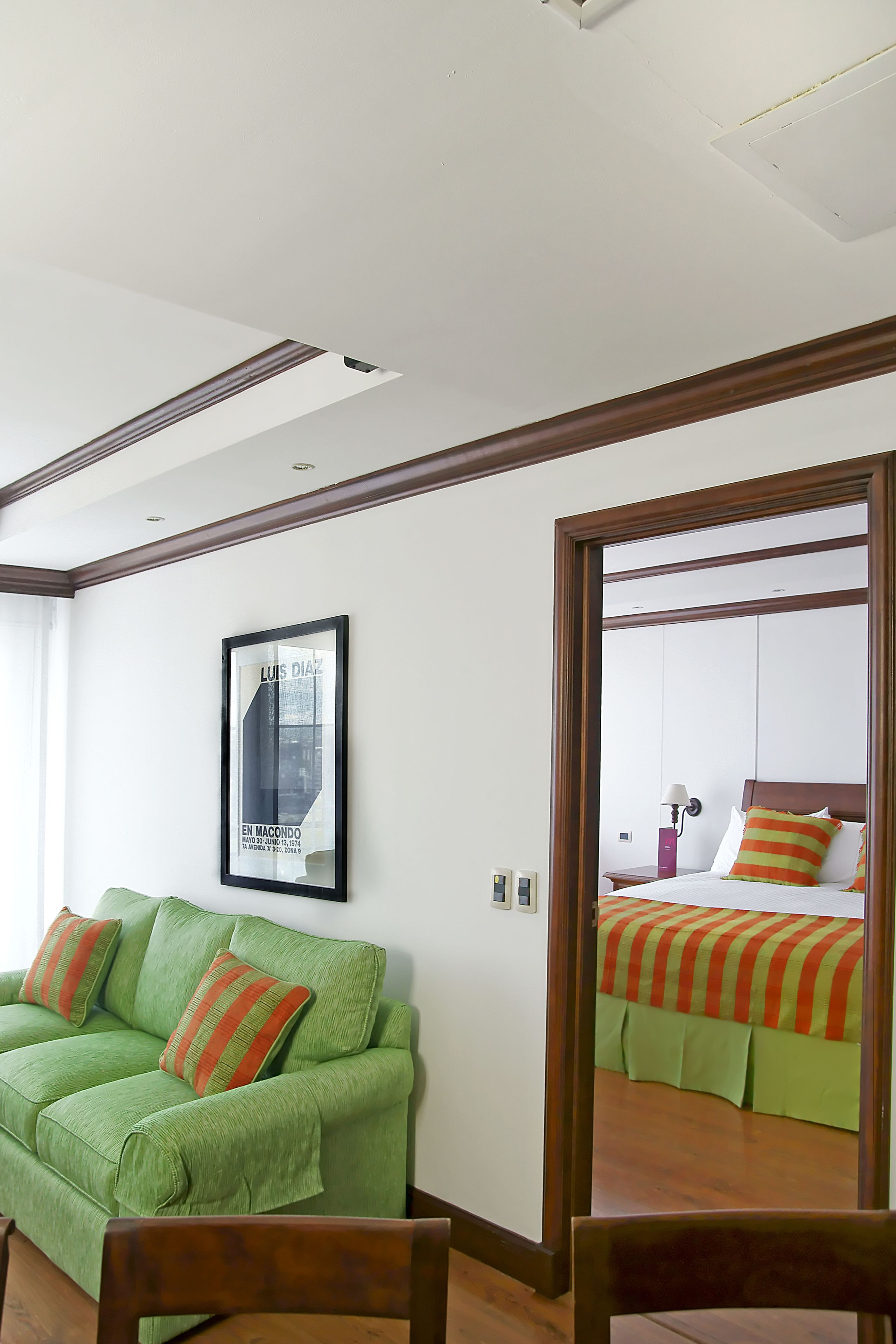 A hotel room with a green sofa, a wooden chair, a framed picture on the wall, and a bed with striped bedding.