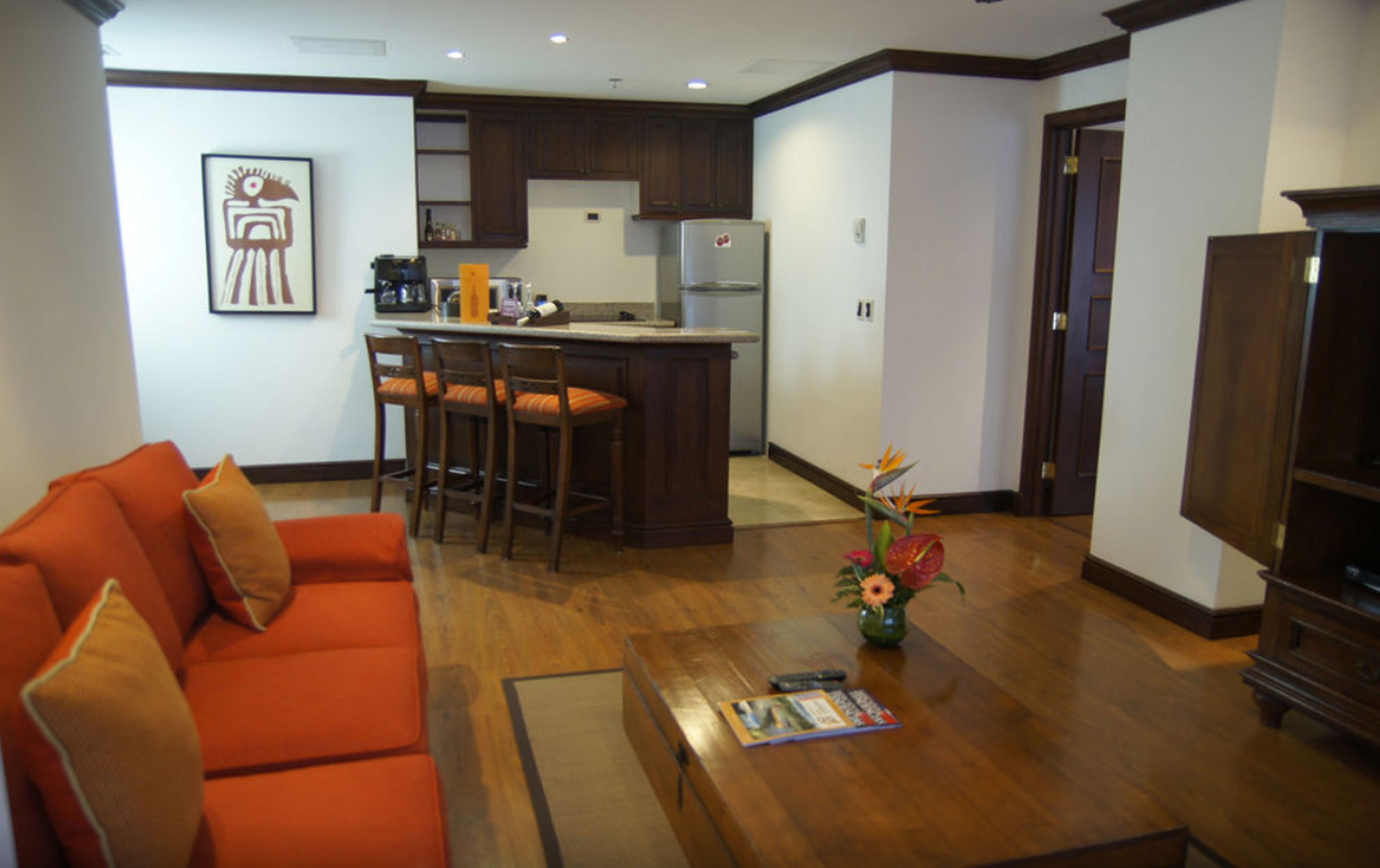 Sala de estar con sofá, una mesa de centro con una revista, y una zona de cocina con refrigerador y taburetes de bar.
