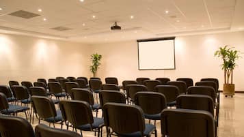 Un salón de conferencias con filas de sillas, una pantalla de proyector y dos plantas en macetas.