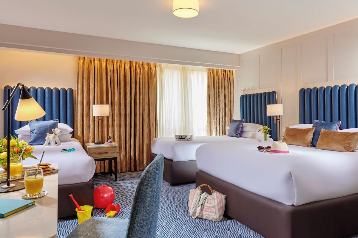 Habitación de hotel con dos camas, un escritorio con lámpara, una silla, una mesita con una bebida y una ventana con cortinas.