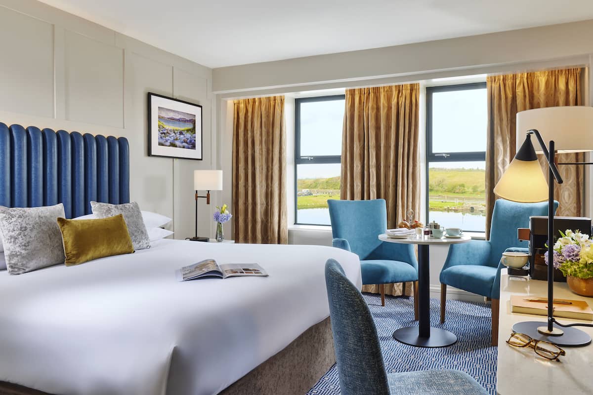 Un cuarto de hotel con una cama grande, dos sillones azules, una mesita redonda y vistas a un paisaje verde por las ventanas.