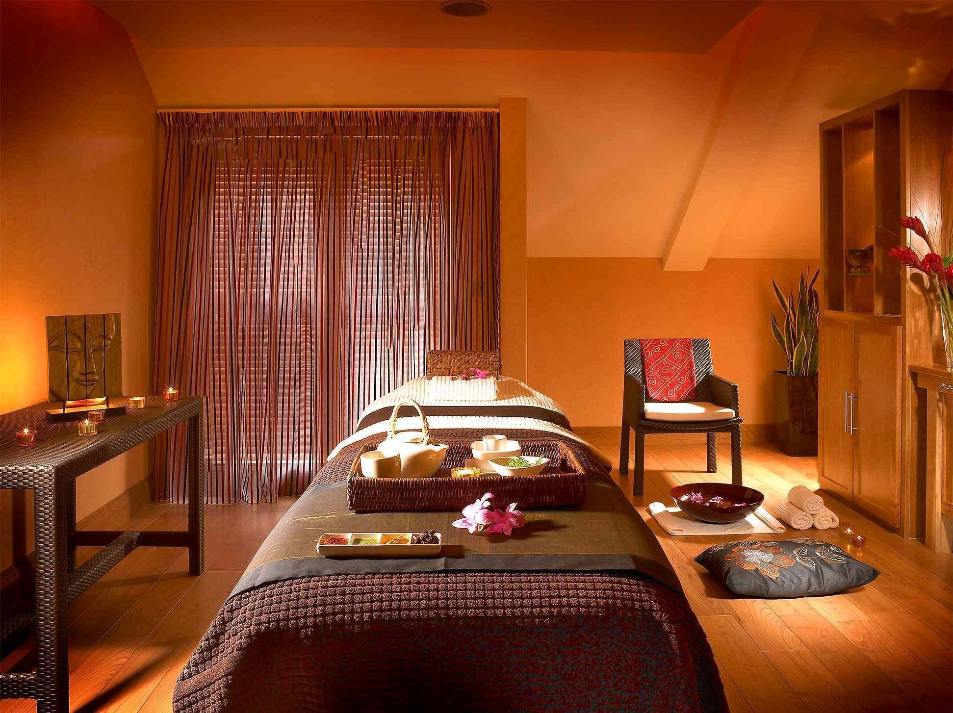 A room with a massage table, a chair, a small table with candles, and a cabinet with plants.