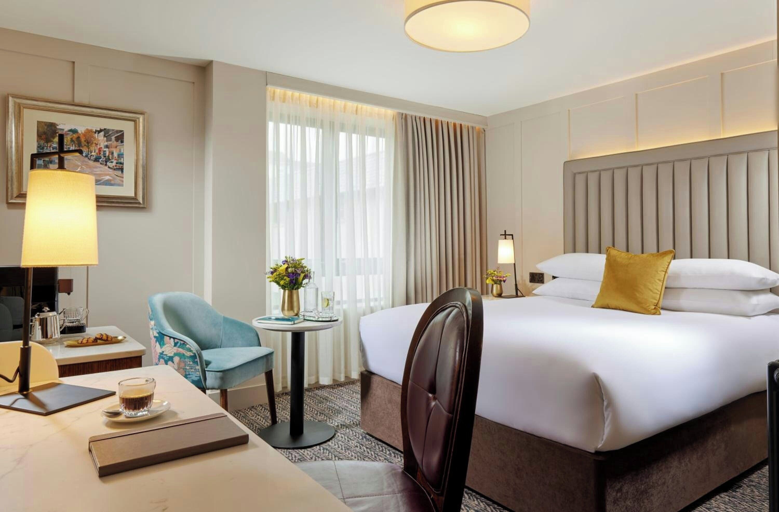 Una habitación de hotel con cama, escritorio, silla, mesa con lámpara y ventana con cortinas.