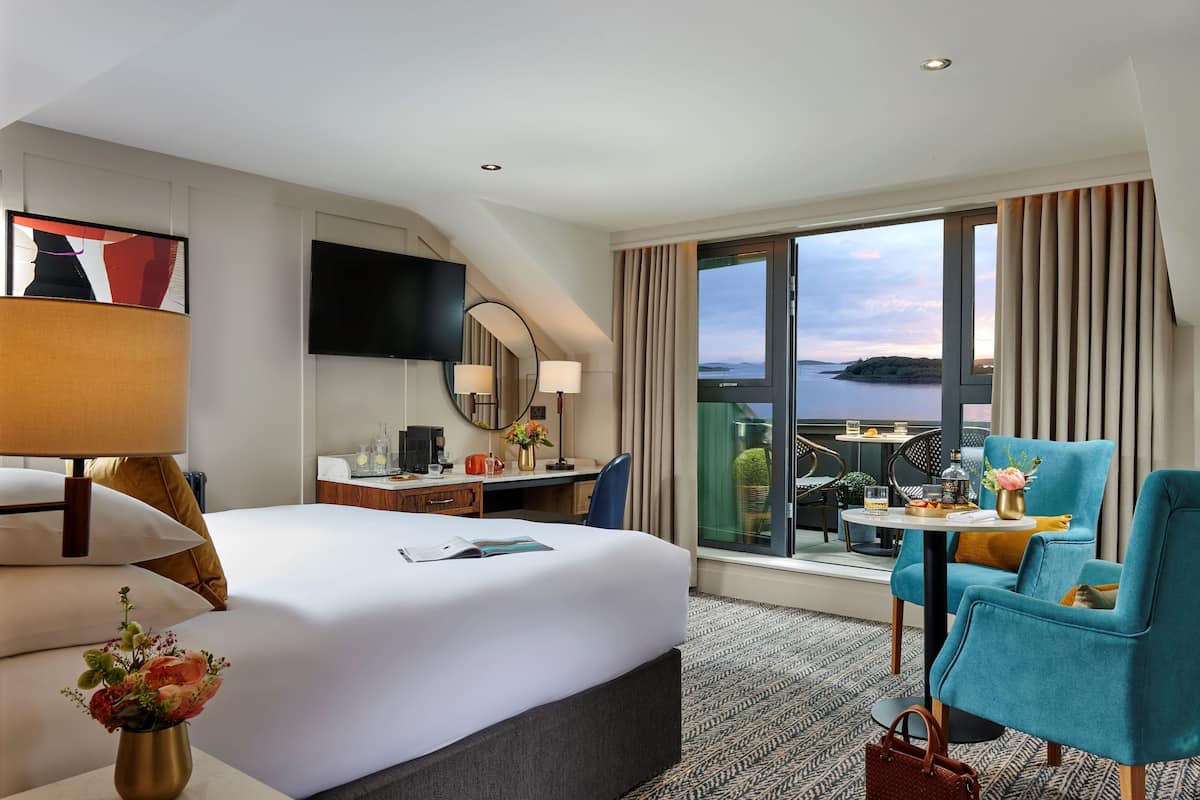 Habitación de hotel con una cama grande, un escritorio, una silla y un balcón con vistas al agua.