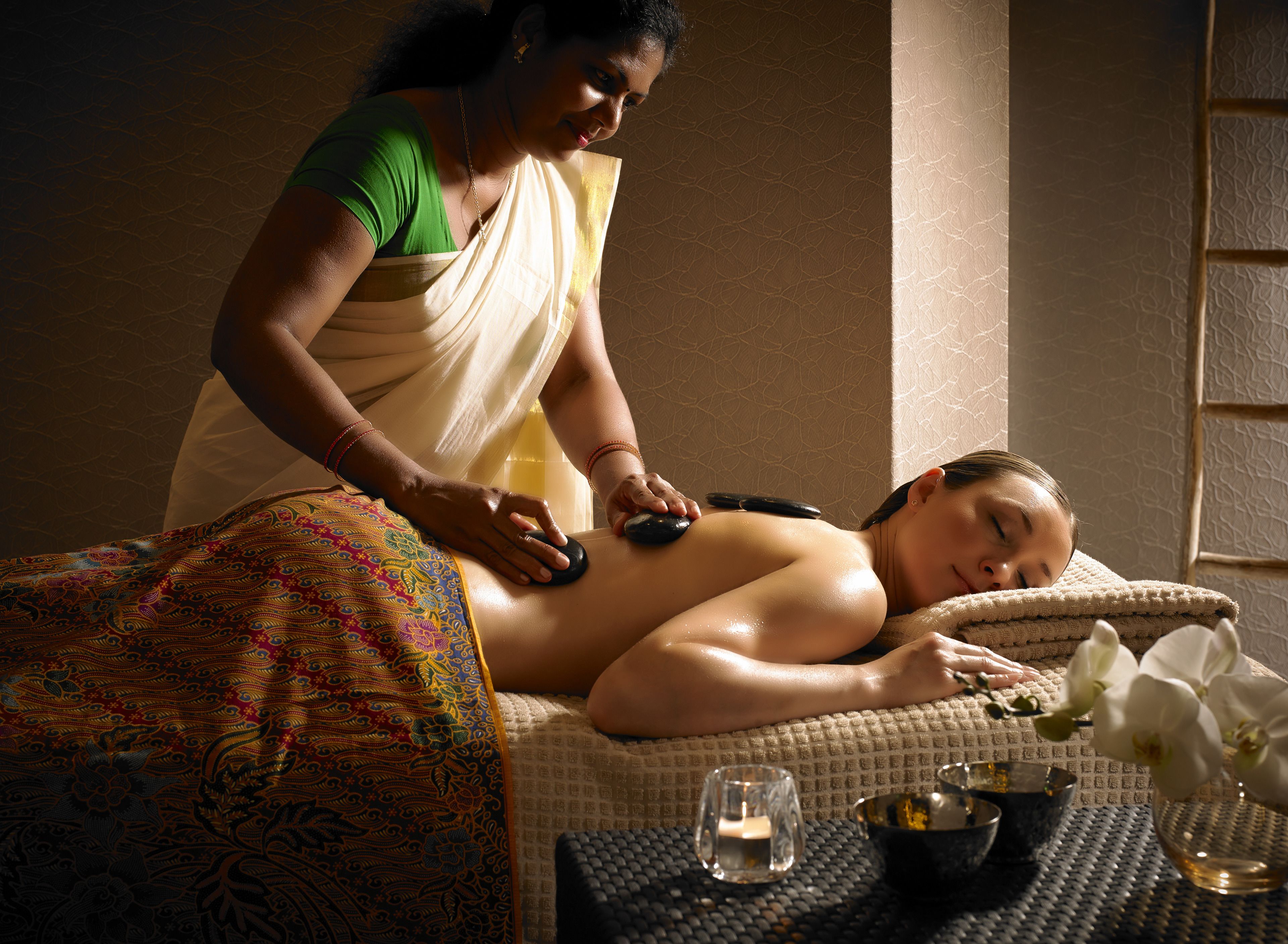 A person receiving a back massage in a room with a patterned bedspread, a small table with a candle, and a vase with flowers.