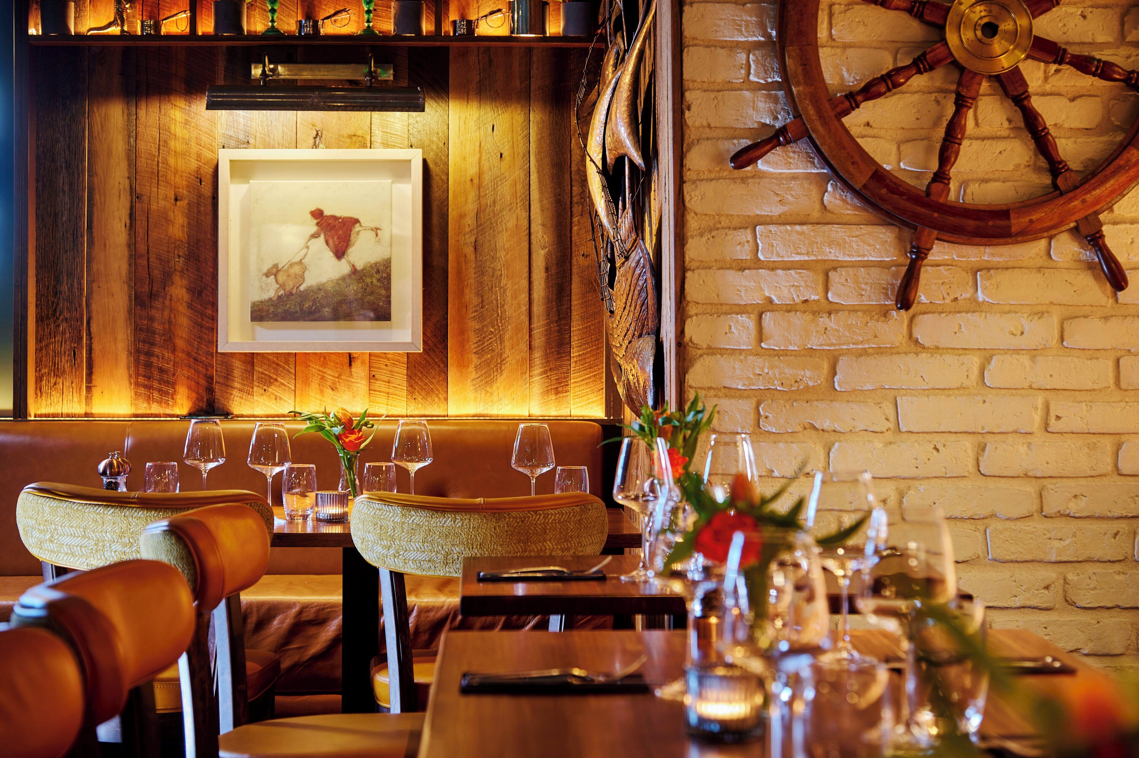 Interior de restaurante con paredes de madera, un cuadro enmarcado, una rueda de barco y una mesa dispuesta con vasos y flores.
