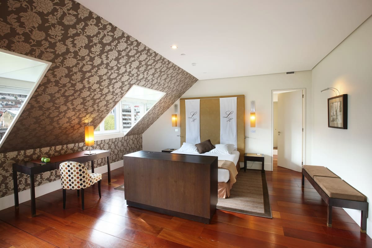 Une chambre moderne avec un plafond en pente, un bureau en bois, une chaise à motifs, un lit avec une tête de lit et un banc.
