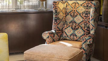 Un sillón estampado con un cojín, una lámpara y una estantería en una habitación con papel tapiz a cuadros y una ventana con cortinas.
