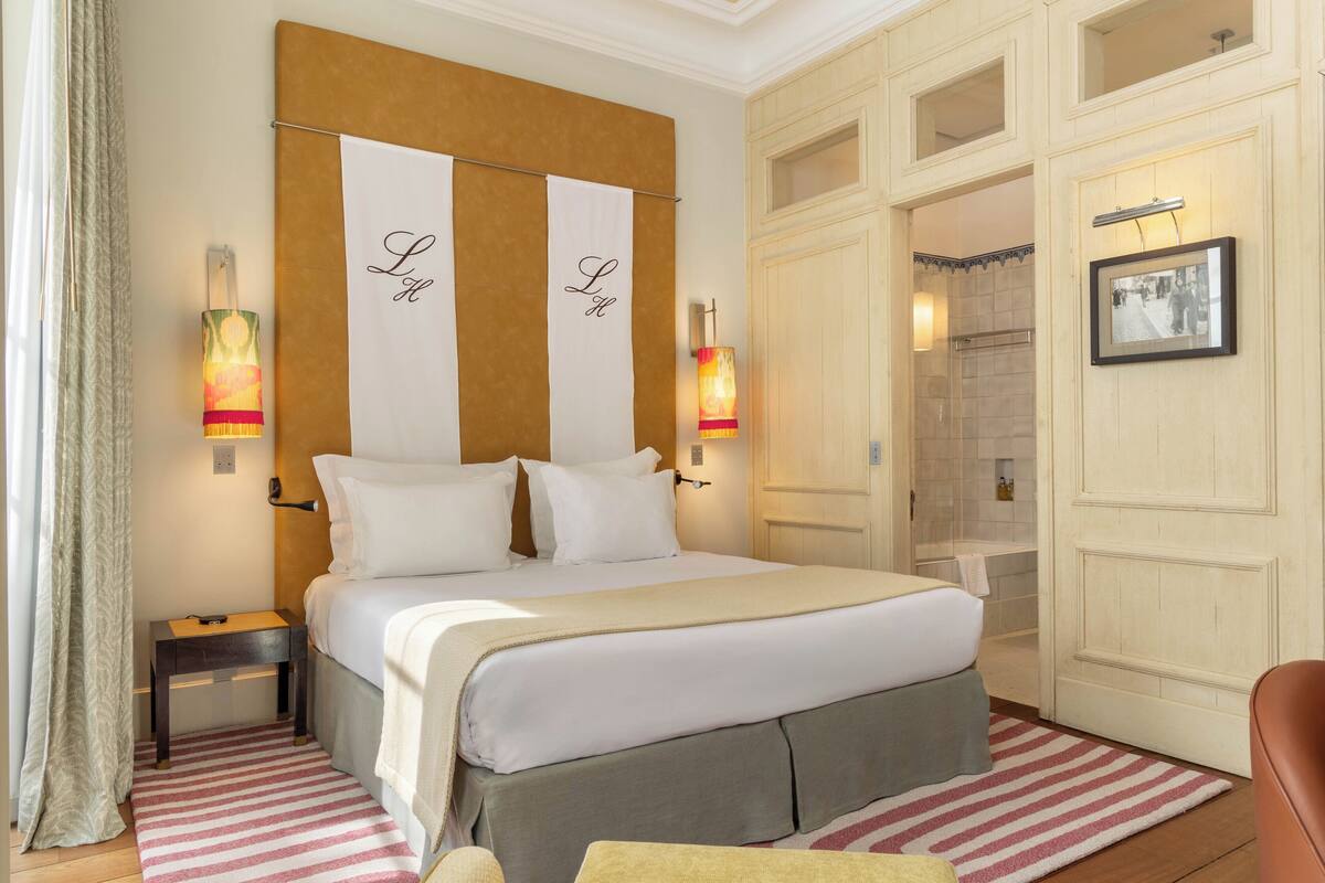 Een hotelkamer met een groot bed, twee bedlampen, een houten hoofdbord, een klein nachtkastje, een gestreept vloerkleed, een bruine lederen fauteuil en een ingelijst schilderij aan de muur.