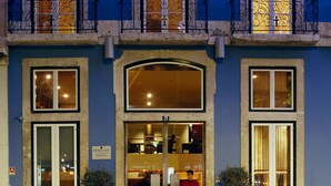 Entrada de un hotel con puertas dobles, un balcón con barandilla de hierro forjado y dos macetas.