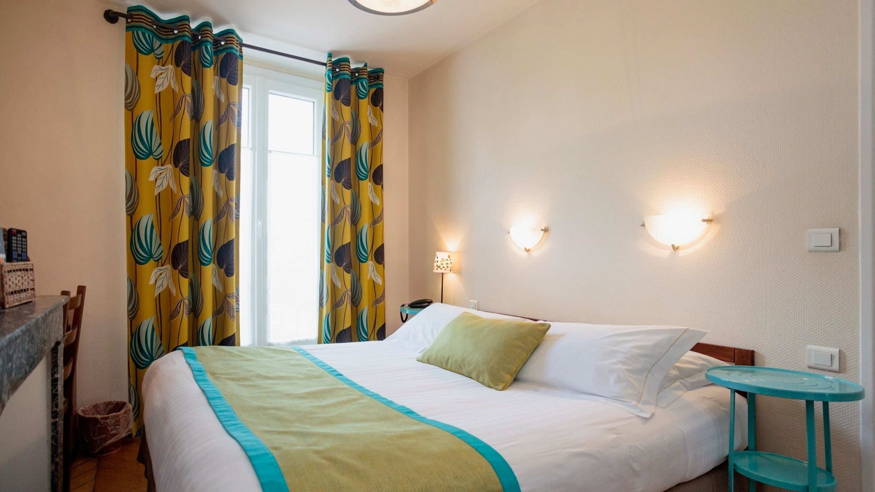 Habitación de hotel con cama, cortinas, lámparas de noche, una mesita y una lámpara fijada a la pared.