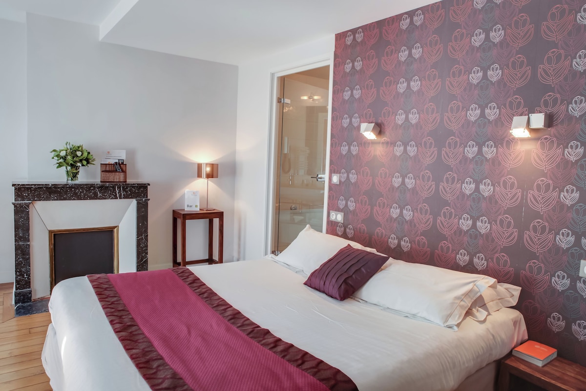 A hotel room with a bed, a fireplace, a nightstand, and a wall with a floral pattern.