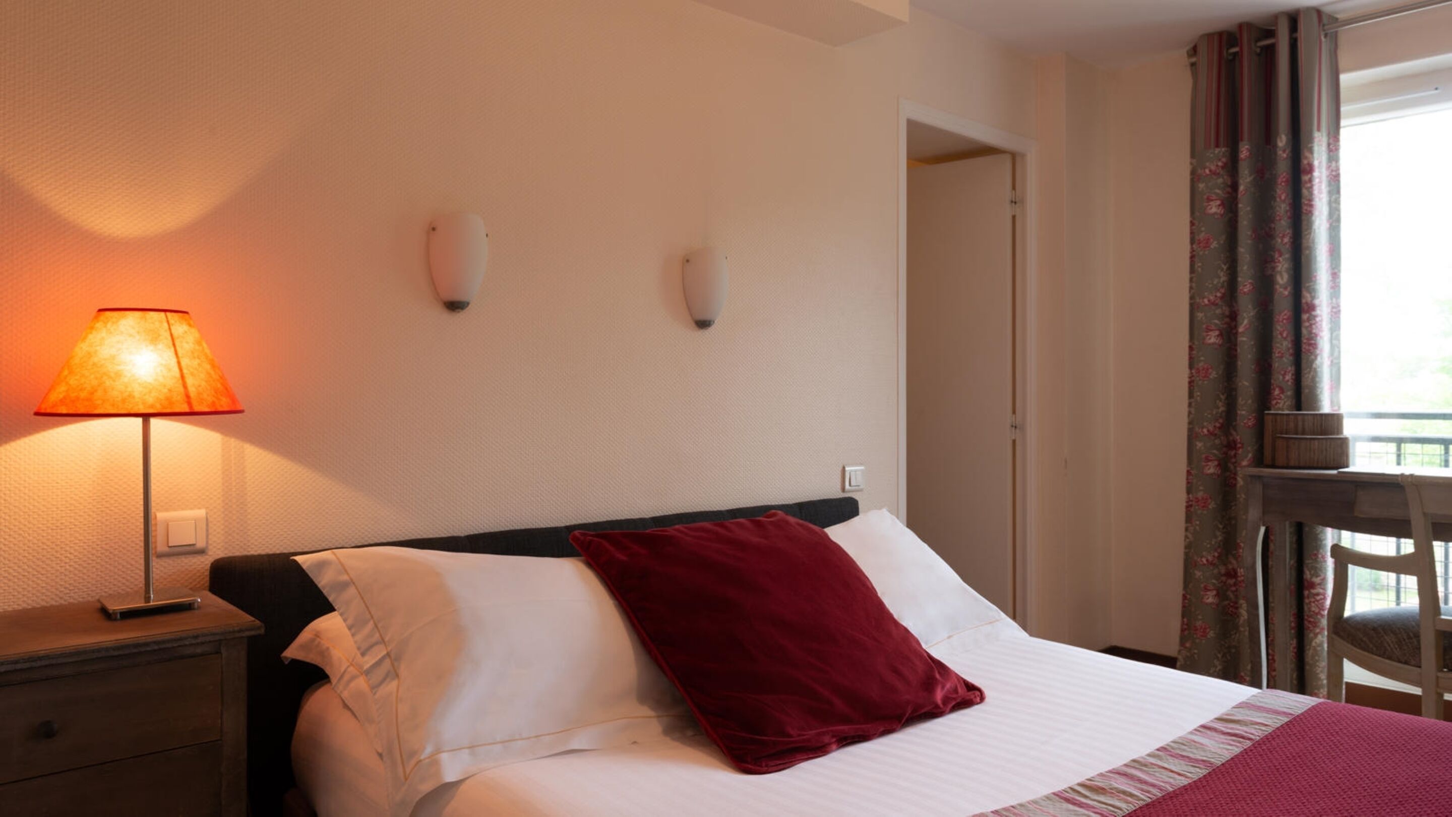 Ein Hotelzimmer mit Bett, Nachttisch, Lampe und Blick ins Freie durch ein Fenster mit Vorhängen.