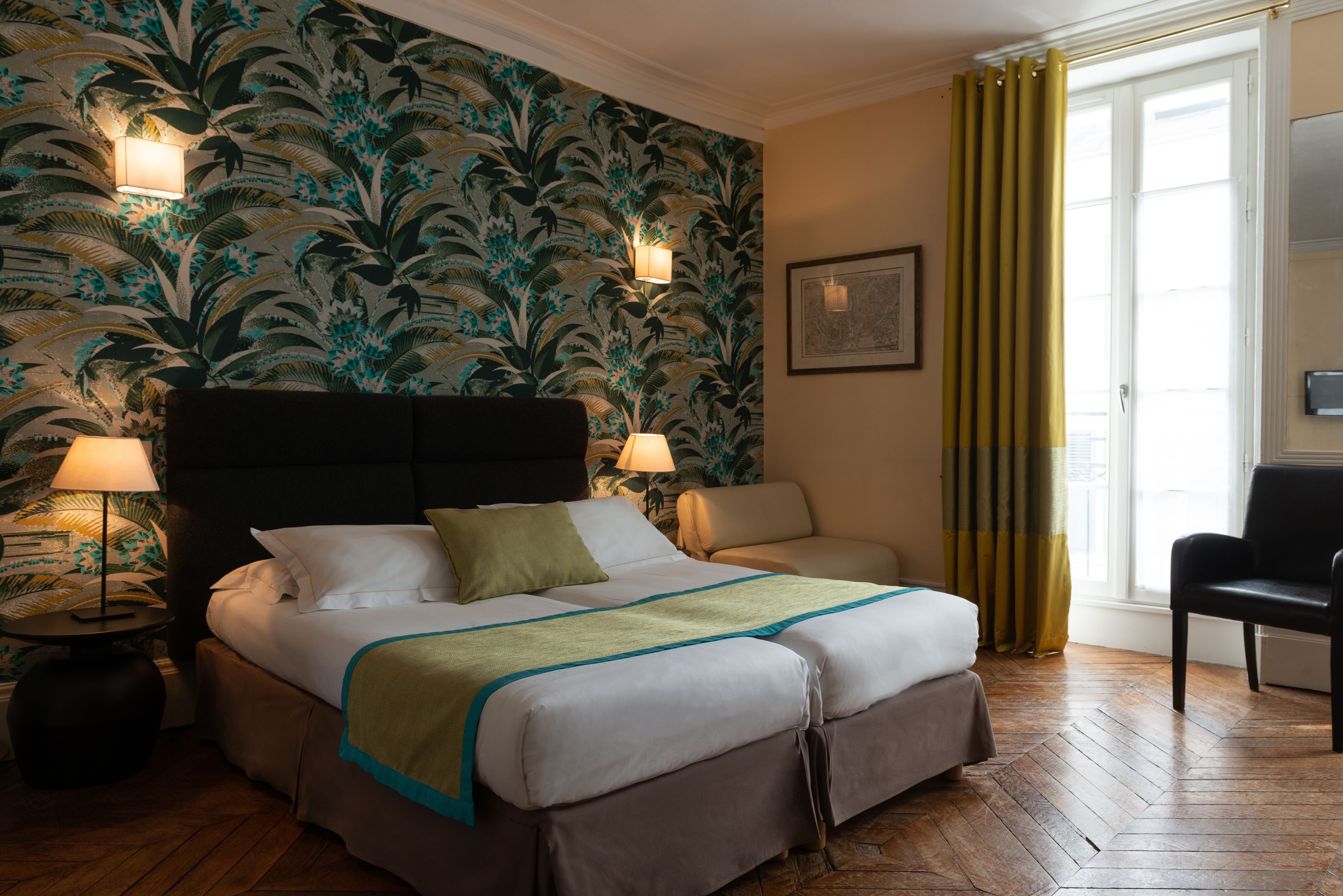 Habitación de hotel con cama, dos lámparas de noche, una silla, ventana con cortinas y pared con estampado floral.