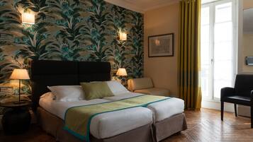 Ein Hotelzimmer mit Bett, zwei Nachttischlampen, einem Sessel, einem Fenster mit Vorhängen und einer Wand mit Blumenmuster.